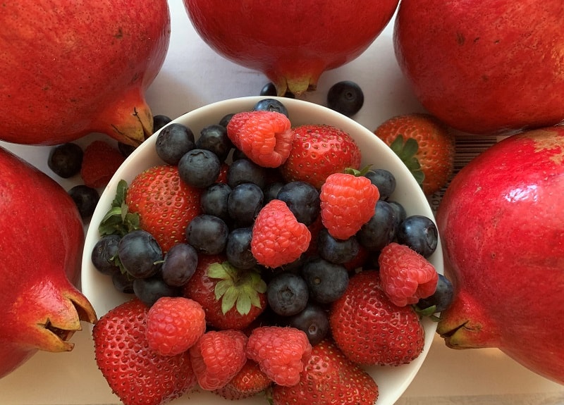 An image of bowl of raspberries and blueberries with pomegranate around the bowl to describe the importance of balanced diet on all organs including skin