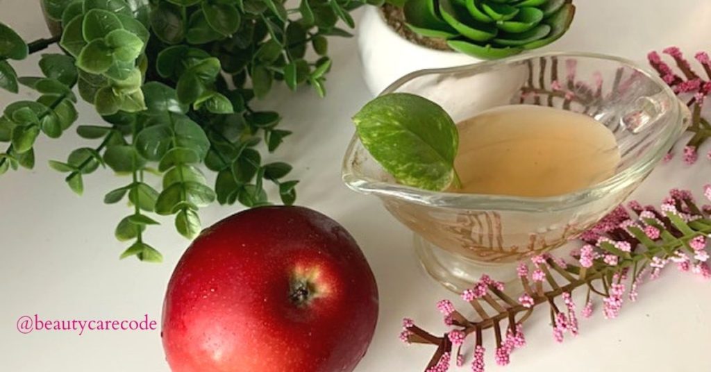 An image of an apple and apple vinegar in a glass container with green plants and pink flower in white background