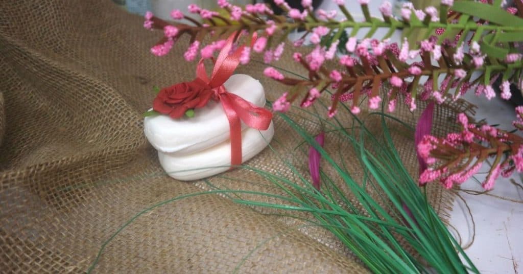 An image of two white soaps with a pink ruban around them and a rose flower on top, and pink flower and herb in the background.